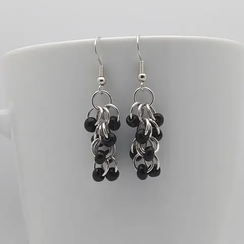 Silver dangly chain mail earrings with black beads hanging on a white mug.