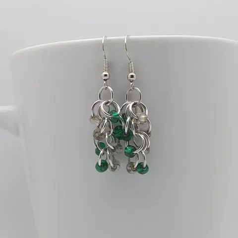 Silver dangly earrings with clear and emerald beads hanging on a white mug.