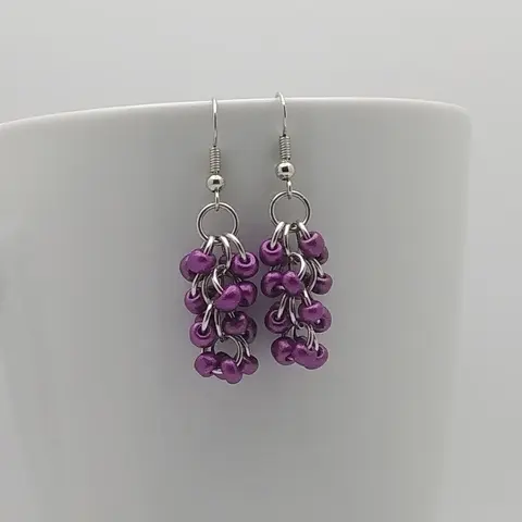 Silver dangly earrings with metallic purple beads hanging on a white mug.