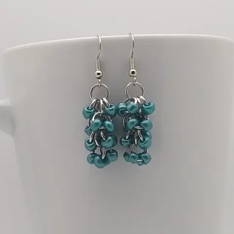 Silver dangly earrings with metallic teal beads hanging on a white mug.