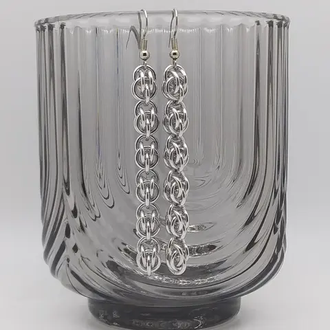 Silver dangly chain mail earrings hanging on a textured cup.