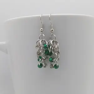 Bright silver aluminum chain mail earrings with clear and emerald beads on a white mug.