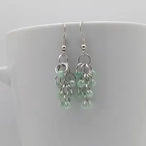 Bright silver aluminum chain mail earrings with green beads on a white mug.