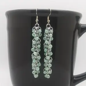 Bright silver aluminum long chain mail earrings with green beads on a black mug.