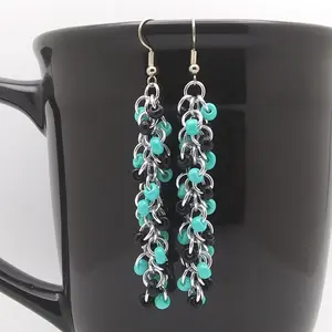 Bright silver aluminum long chain mail earrings with black and turquoise beads on a black mug.